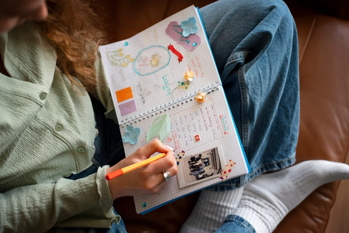 Person seated, writing in a decorated notebook with stickers and drawings, wearing jeans and a green cardigan.