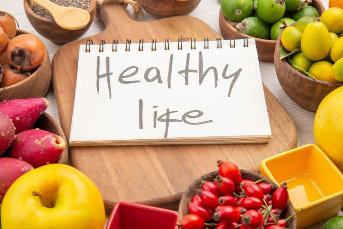 Notebook labeled "Healthy Life" surrounded by bowls of fresh fruits and wooden utensils on a table.