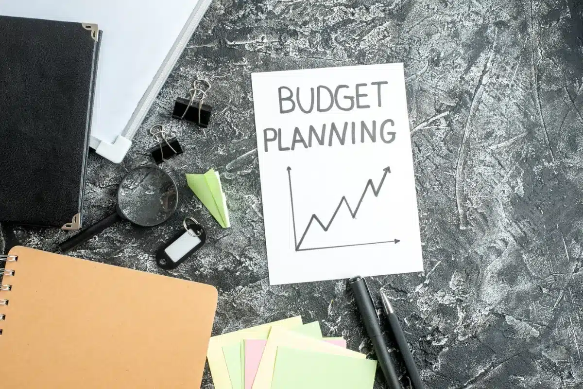 A desk with a Budget Planning chart, office supplies, and notebooks on a textured grey background.