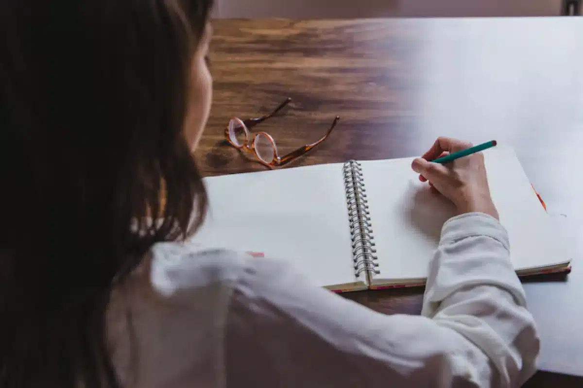 Woman writing in a notebook at a wooden table with glasses beside.
