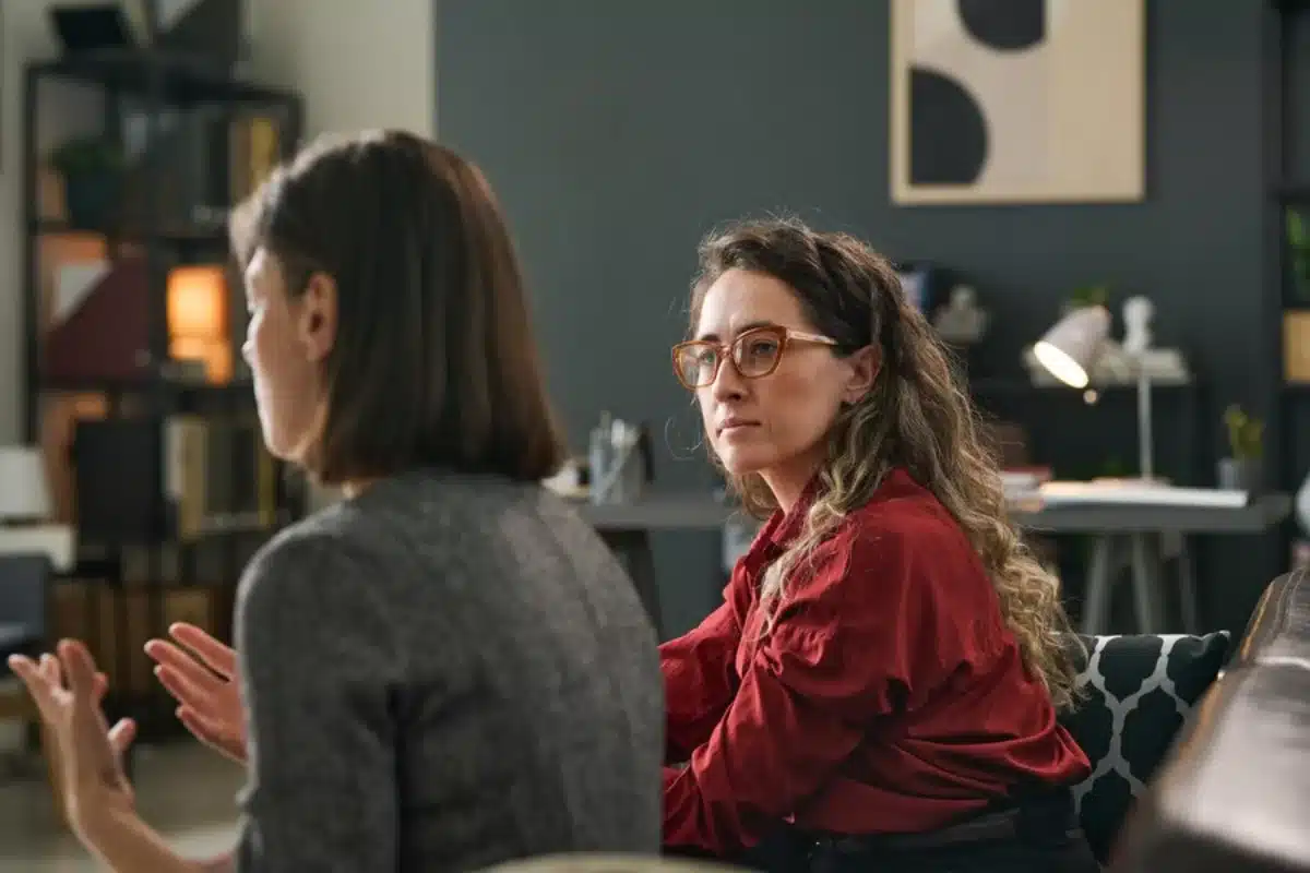 Two women in conversation, one in a red shirt and glasses listening intently in a modern, dimly-lit living room.