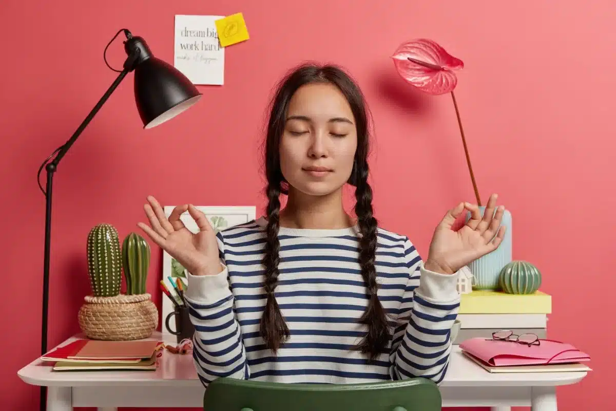 Young woman meditating at a colourful study desk with plants and lamp, eyes closed, peace sign with fingers.