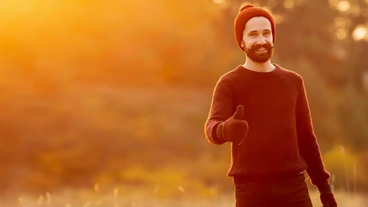 Man in a burgundy jumper and orange beanie giving a thumbs-up in a sunlit autumn field.