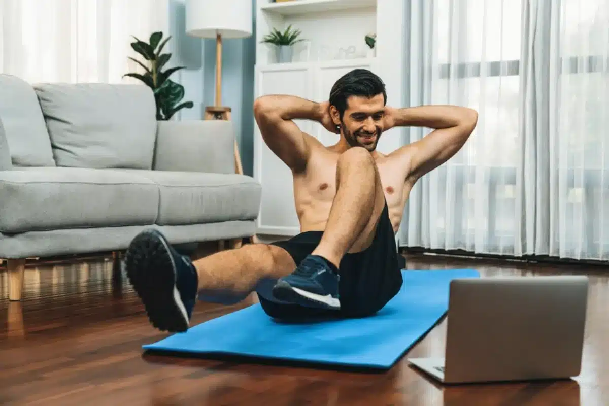 Man doing sit-ups at home on a blue mat, watching a laptop, with sofa and plants behind.